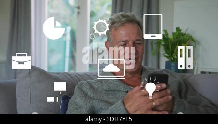 Holding smartphone, senior man wearing grey cardigan sitting on sofa in home, showing tech icons Stock Photo