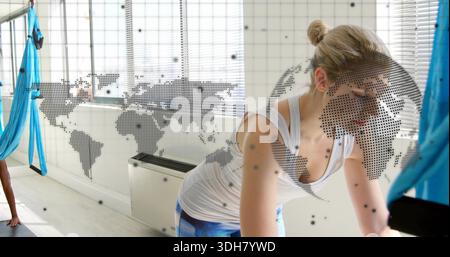 Leaning blonde woman wearing white tank and blue leggings in studio with silk hammock and world-map Stock Photo