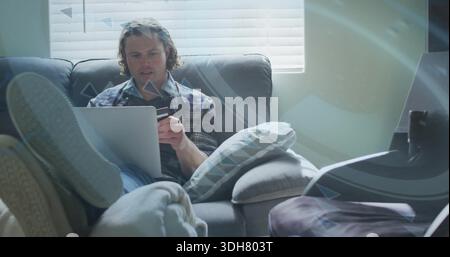 Holding smartphone, man in plaid shirt checking messages on gray couch at home, laptop on lap Stock Photo