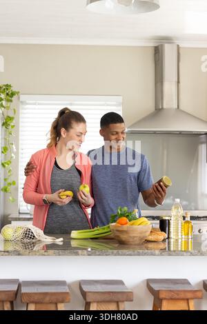 Couple standing at kitchen island, woman in coral jacket holding apples, man examining jar Stock Photo