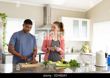 Diverse couple chopping cucumber on wooden cutting board at marble island in modern kitchen Stock Photo