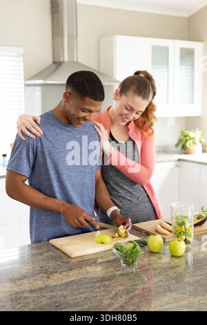 Diverse couple preparing fruit at kitchen island, man in blue tee slicing apple on cutting board Stock Photo