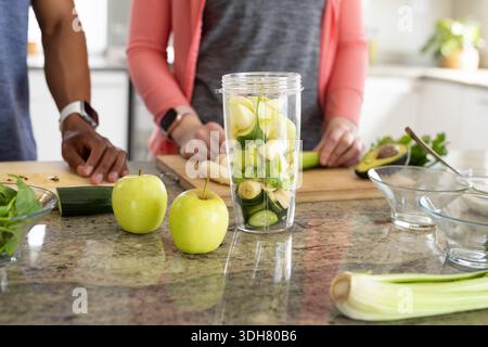 Diverse couple preparing green smoothie at kitchen counter with blender wearing smartwatches Stock Photo