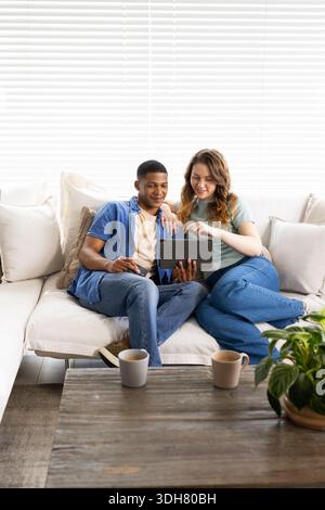 Diverse couple sitting on light sectional sofa sharing tablet, pointing at screen, casual clothing Stock Photo