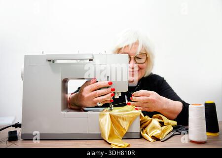 Senior woman is sewing golden fabric on a sewing machine, creating a garment Stock Photo