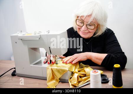 Senior woman enjoying creative hobby, using a sewing machine to work on golden fabric Stock Photo