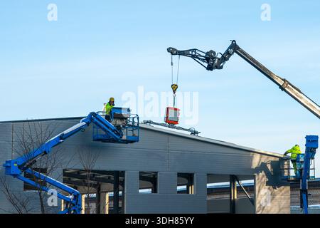 Construction workers install rooftop equipment using modern crane and boom lifts on industrial building under clear blue sky Stock Photo