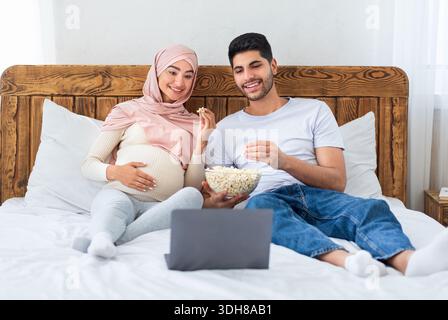 Leisure time. Relaxed arab man and pregnant wife watching film on laptop computer, eating popcorn, sitting on bed Stock Photo