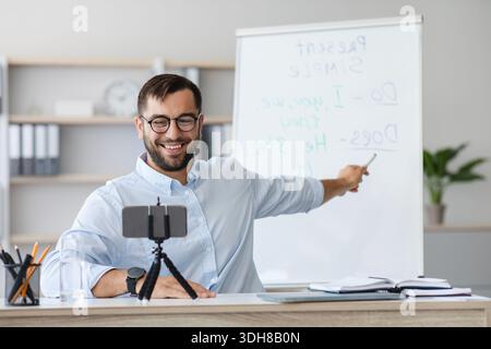 Cheerful adult european male teacher with beard in glasses shows to blackboard and have video call Stock Photo