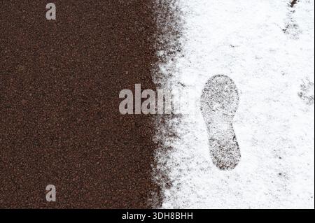 lonely footprint beside dark surface, horizontal composition showing brown textured terrain meeting snow, crisp sole detail and subtle grain, suggesti Stock Photo