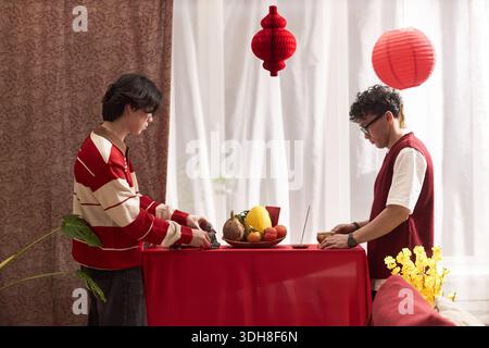 Two Asian young adults preparing traditional holiday offerings on red table with fruit centerpiece, standing under red lantern decorations during Tet celebration indoors Stock Photo