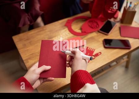 Asian young adult writing on red envelope with pen during Tet holiday, sitting at wooden table with other red envelopes, smartphones and festive decorations visible in background Stock Photo