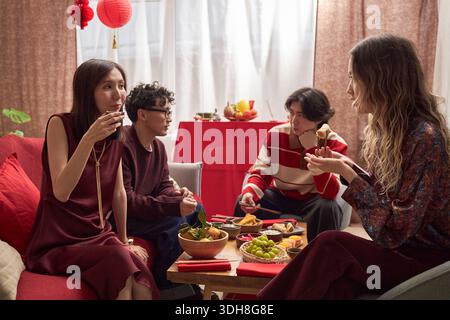 Group of young adults including Asian woman, Asian man, Caucasian man and Caucasian woman sitting together enjoying traditional food and drinks during Tet holiday celebration indoors Stock Photo