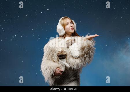 Studio portrait of woman in fluffy coat and earmuffs catching snowflakes on blue background for winter fashion and cold weather ads Stock Photo