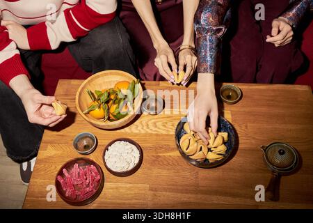 Three young adult Caucasian and Asian women sitting together reaching for fortune cookies and traditional snacks on wooden table, celebrating Tet holiday with festive treats Stock Photo