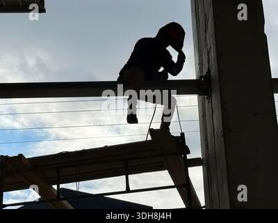 Silhouette of a welder working on a high-altitude steel beam at a construction site. Sparks fly against a bright sky. Industrial labor and safety Stock Photo