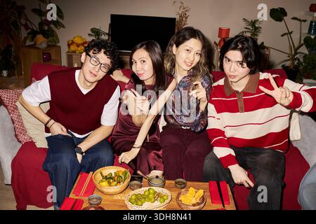 Portrait of group of young adult Asian men and women sitting together on sofa, smiling and making peace signs while celebrating Tet holiday with traditional food on table in living room Stock Photo