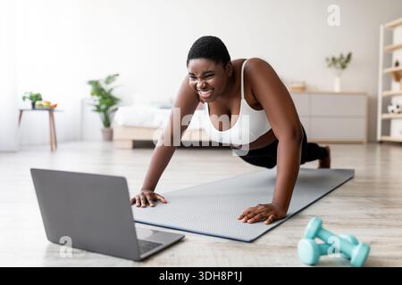 Plus size Afro woman standing in plank pose or doing push ups near laptop, working out to online sports tutorial at home Stock Photo