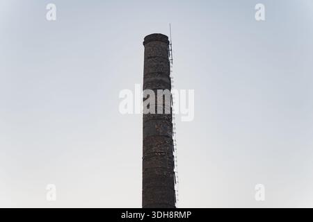 Tall industrial brick chimney with metal ladder rising against clear sky showing former factory infrastructure and vertical industrial architecture Stock Photo