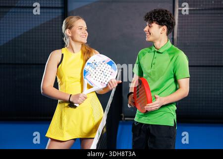 Happy man and woman with padel rackets talking by the net on indoor court during lesson practice and club lifestyle activity Stock Photo