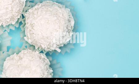 Three white coconut balls are sitting on top of a blue background. The coconut balls are in paper cups and are arranged in a row Stock Photo