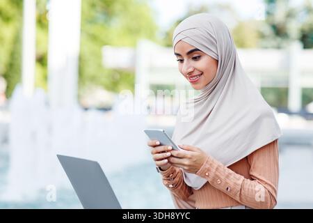 Busy smiling cute millennial arab muslim lady in hijab typing on smartphone, use laptop Stock Photo