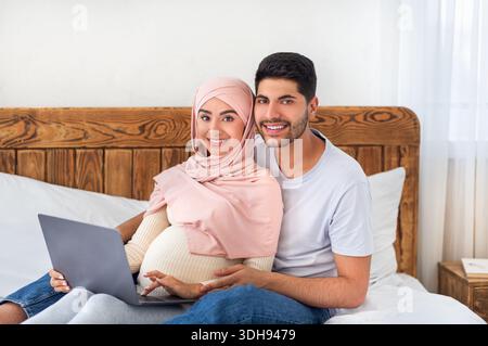 Loving pregnant muslim couple using laptop together, surfing internet and spending time together, sitting on bed Stock Photo