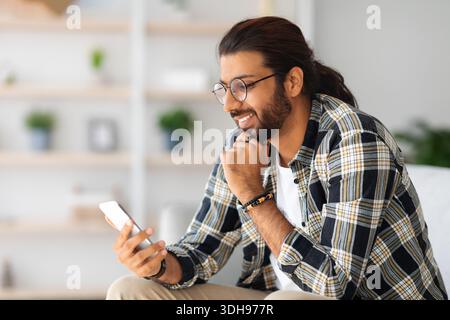 Cheerful long-haired middle-eastern guy hipster sitting on couch, using smartphone Stock Photo