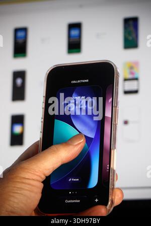 Person holding an iPhone smartphone customizing lock screen display with various wallpaper options shown on a computer screen in the background Stock Photo