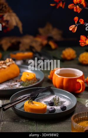 Plating an orange pumpkin roll dessert with fresh blueberries and thyme sprigs on a dark green plate, complementing a pumpkin shaped mug of hot tea an Stock Photo