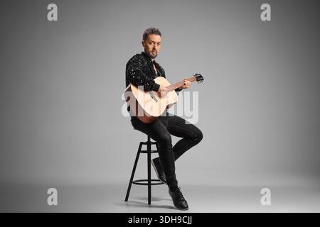 Male musician sitting on stool with an acoustic guitar over gray background Stock Photo