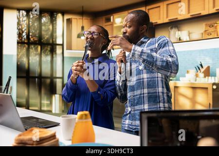 Goofy cheerful people singing together with tools as microphones in the kitchen, black couple showing love and playful energy, fooling around and having fun with music. Positive relations. Stock Photo