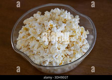 A bowl of freshly popped microwave popcorn in a clear bowl placed on a brown wooden table top. Stock Photo