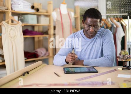 Male tailor draws sketch of future clothing on an electronic tablet. Working with modern gadgets in sewing workshop Stock Photo