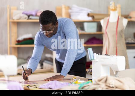 Male tailor draws sketch of future clothing on paper using patterns in sewing workshop Stock Photo