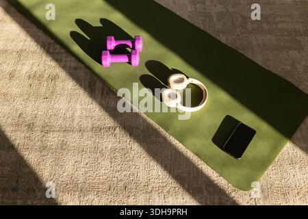 Green yoga mat lying diagonal on beige carpet, sunlight casting shadows over weights headset phone Stock Photo