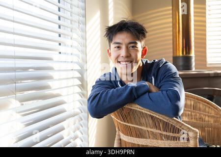 Adult Asian man sitting on wicker chair by window with white horizontal blinds, wearing blue hoodie Stock Photo