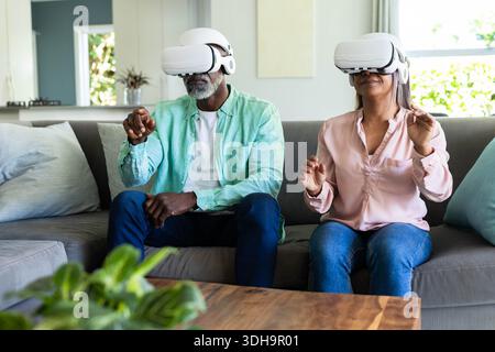 Senior couple exploring virtual world on gray sofa in living room with white VR headsets Stock Photo
