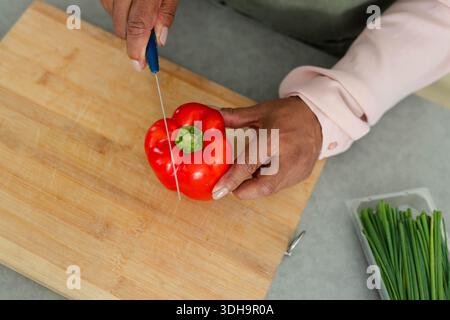 Red bell pepper is being sliced with blue-handled paring knife on gray countertop, chives nearby Stock Photo