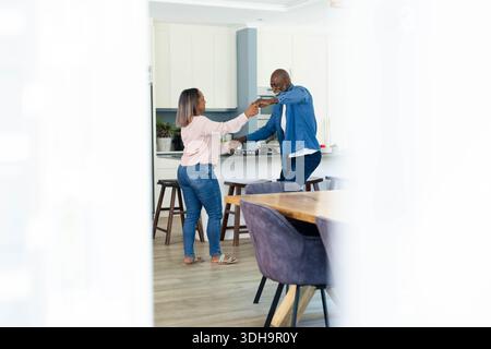 Diverse mature couple holding hands and dancing in modern home kitchen near potted plant Stock Photo
