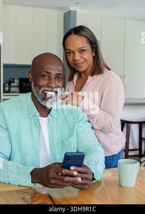 Diverse couple sitting in kitchen, man in green shirt holding blue phone, woman resting on shoulder Stock Photo