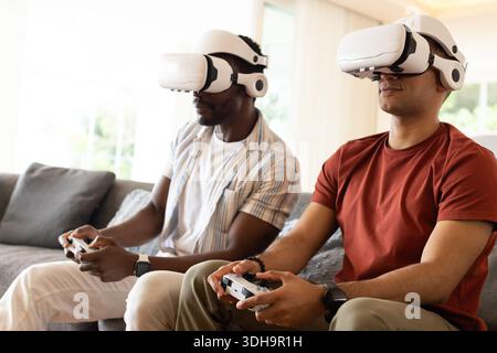 Diverse male friends sitting on gray sofa wearing white VR headsets and holding controllers Stock Photo