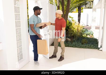 Two adult men delivery worker in polo passing pizza boxes to resident in beanie at doorway Stock Photo