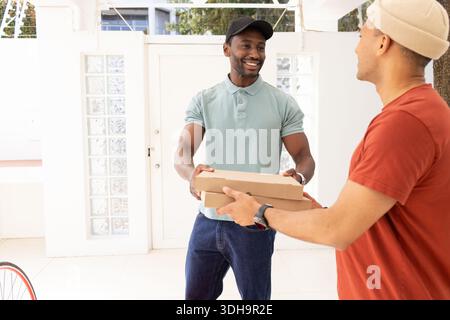 Two adult men exchanging flat cardboard food boxes at home entryway by white door and bicycle Stock Photo