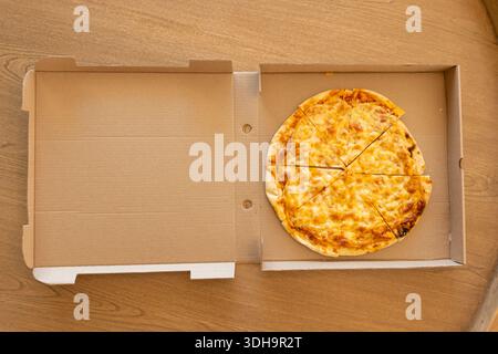 Round cheese pizza is resting in open corrugated box on wooden tabletop, showing charred crust Stock Photo