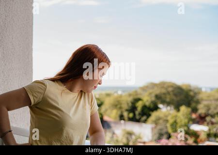 Young red-haired woman leaning on a balcony railing looking down over a residential city view, thoughtful mood. Stock Photo