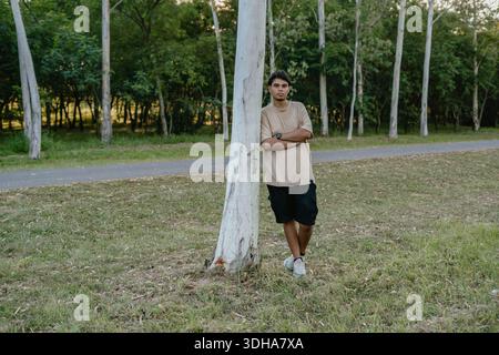 Young man leaning against a tree with arms crossed in a public park, relaxed outdoor portrait. Stock Photo