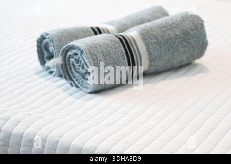 Rolled gray towels neatly arranged on white quilted bedspread in a bright minimalist bedroom. Stock Photo