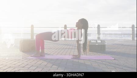 Holding reverse plank, woman exercising on seaside promenade with pink mat, crop top, leggings Stock Photo