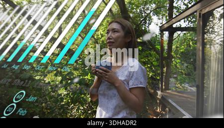 Standing midadult-Chinese woman in top holding ceramic mug gazing at trees on deck with AR chart Stock Photo